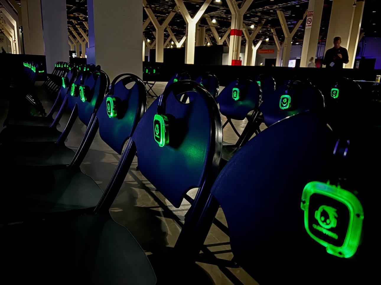Many chairs set with green glowing head phones which are on a local radio broadcast such that the presenter on stage's mic is played through many headphone instead of stage speakers. This permitted many presentations to occur near one another in a shared space without noise from each other. However, the head phones themselves transmitted noise from the microphone and audio mixing deck into everyone's ears.