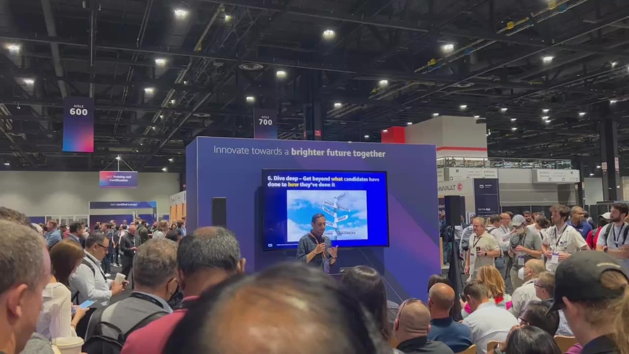 Too many people crowd around one person with a microphone speaking a public vendor hall. There is no sense of personal distance. Above the TV with the presentation, it reads 'Innovate towards a brighter future together'